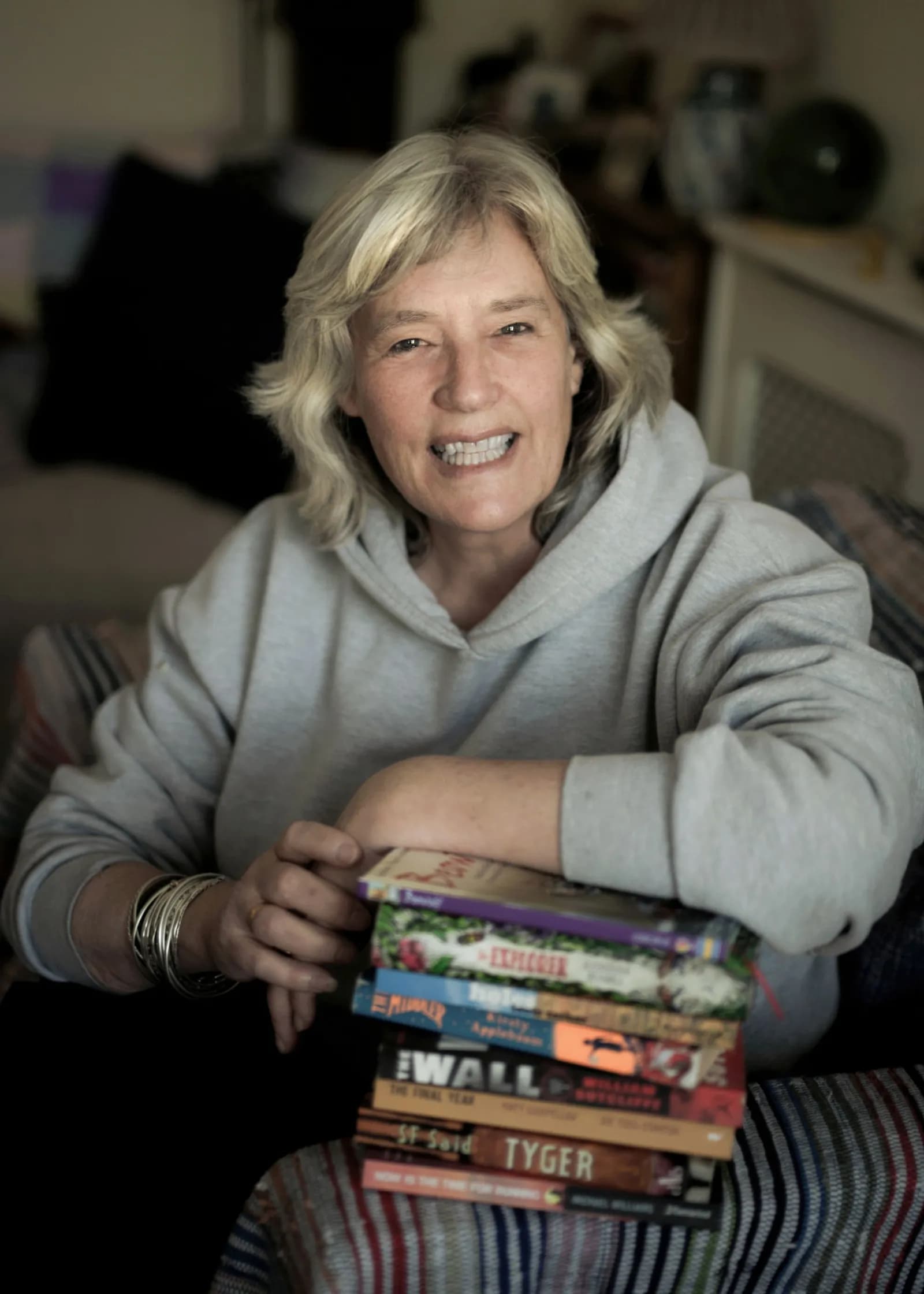 A woman with blonde hair, wearing a gray hoodie, smiles while resting her arm on a stack of books.