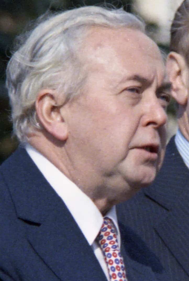 A fair-skinned man with receding gray hair, wearing a dark suit, white collared shirt, and a red, white, and blue patterned tie, looks slightly to the right.
