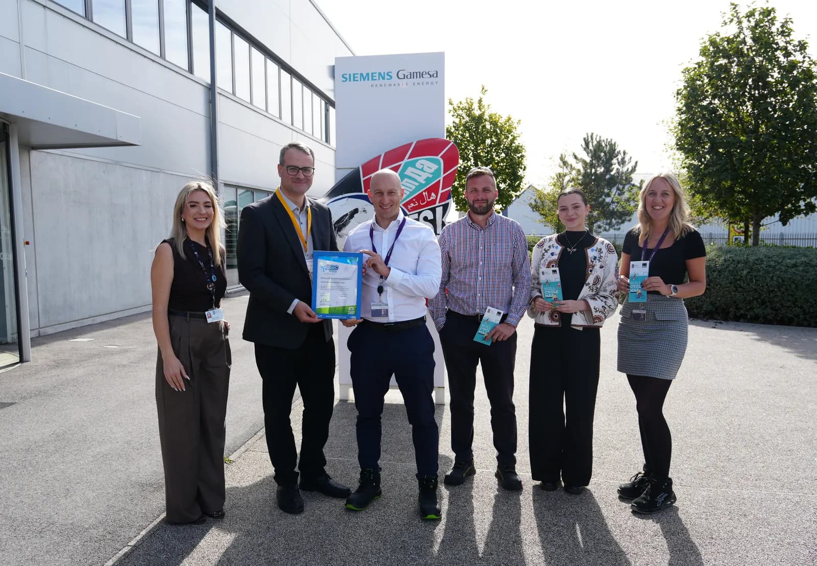 Six people, three men and three women, stand outdoors in front of a building with a large "Siemens Gamesa" sign, with two of the men holding a framed certificate.