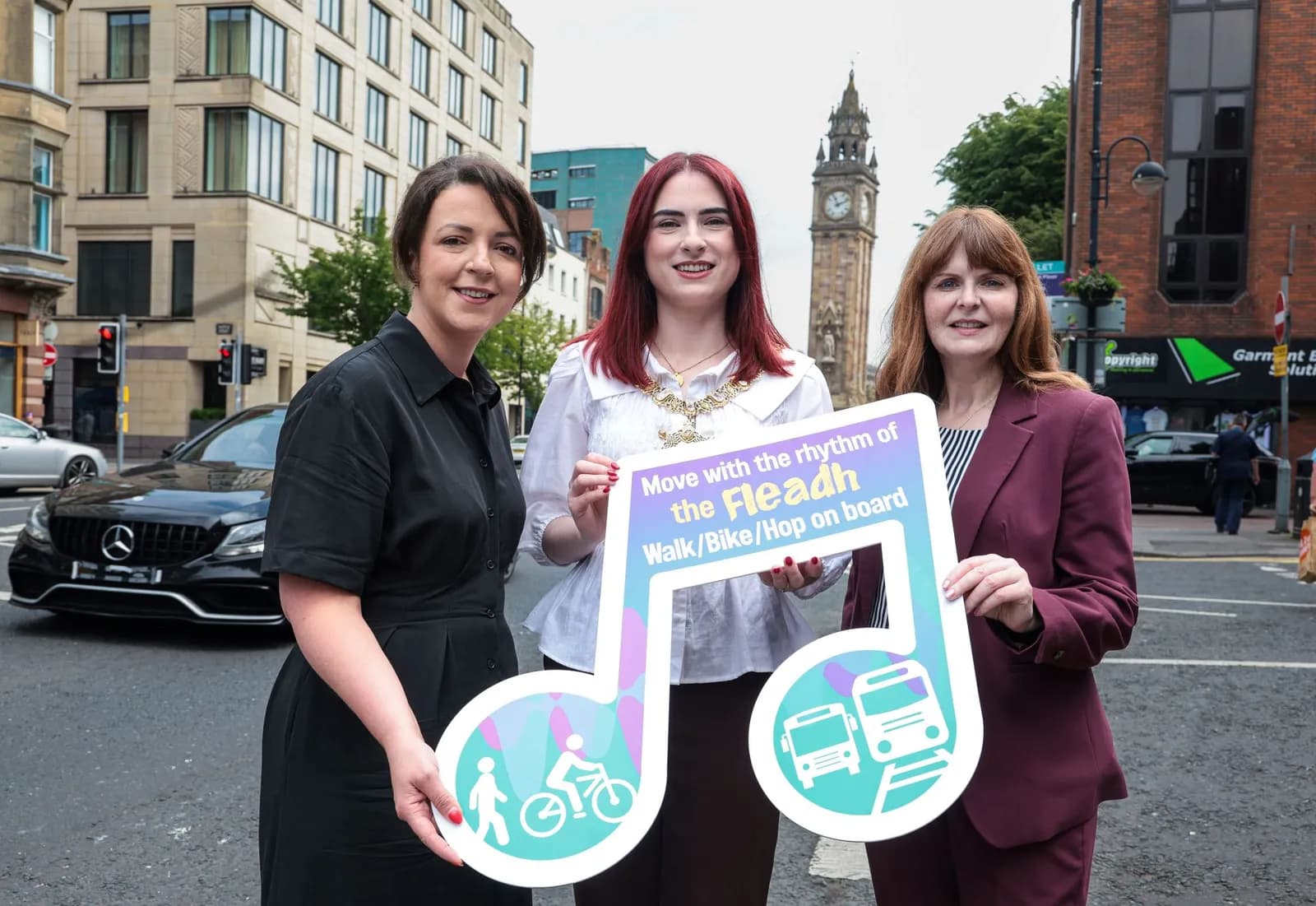 Three women standing on a city street, holding a music note-shaped sign that reads "Move with the rhythm of the Fleadh Walk/Bike/Hop on board".