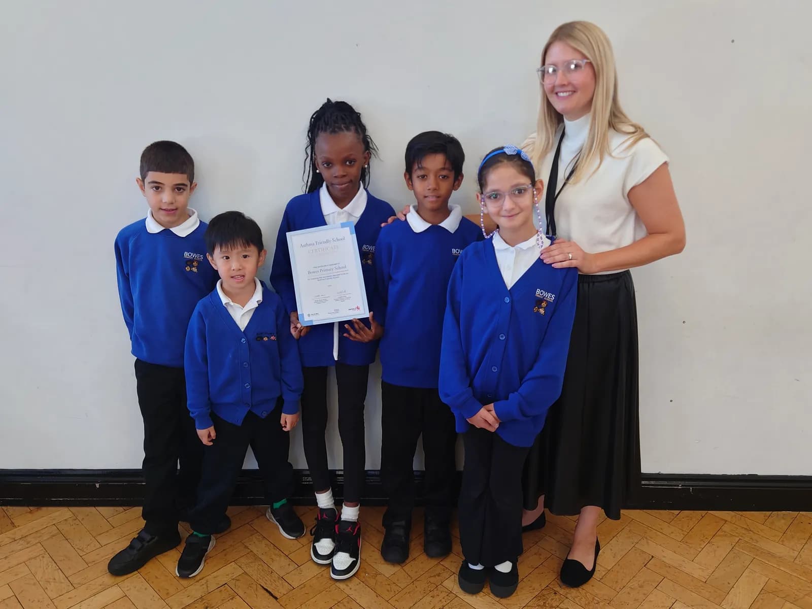 A woman and five children, dressed in blue school uniforms, pose for a photo with a certificate in a room with a white wall and wooden floor.