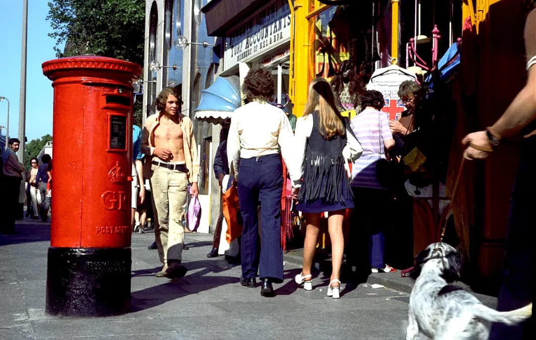 A red pillar box stands on a sunny street next to people walking by, entering and exiting shops, and a dog on a leash in the bottom right corner.