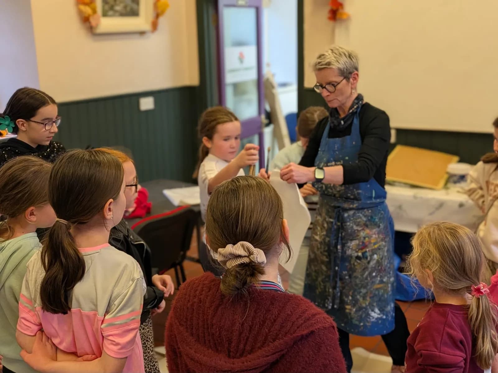 A female art instructor wearing a paint-splattered apron demonstrates a craft to a group of young girls in a classroom setting.