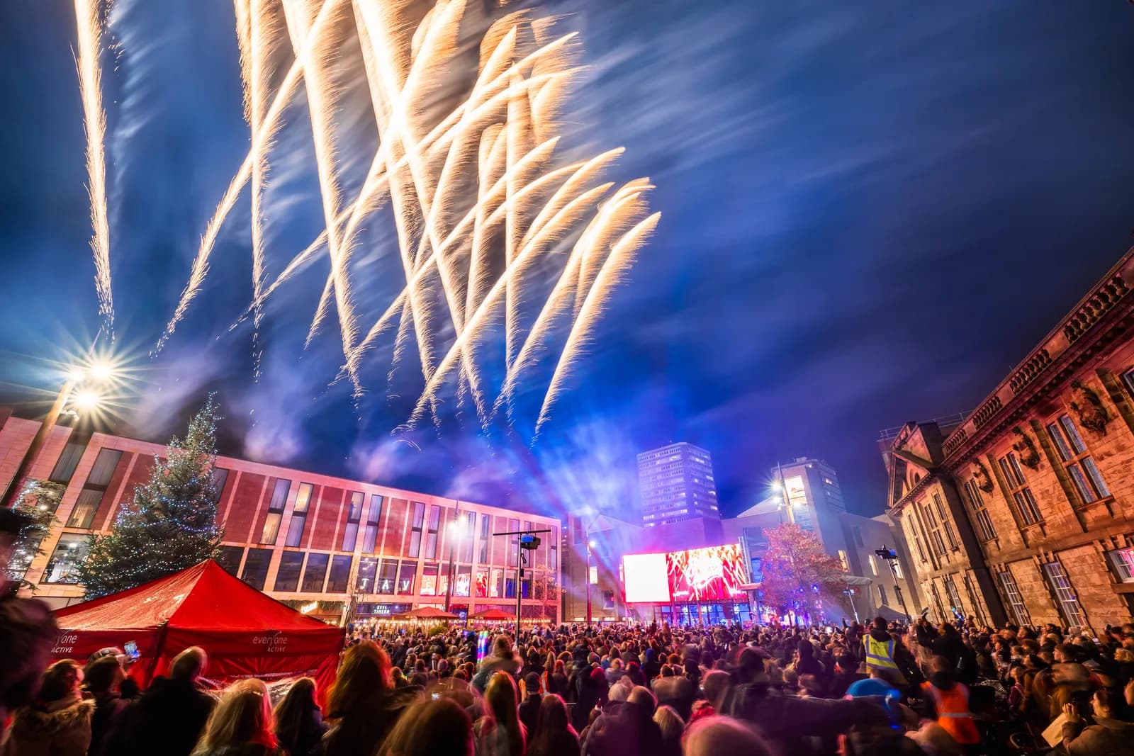 Spectacular golden fireworks burst over a city crowd and buildings, with a lit Christmas tree and red tent visible on the left.