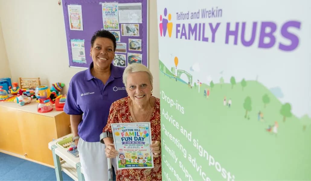 Two women, one in a purple polo shirt and the other in a patterned blouse holding a flyer, stand smiling next to a Family Hubs banner.