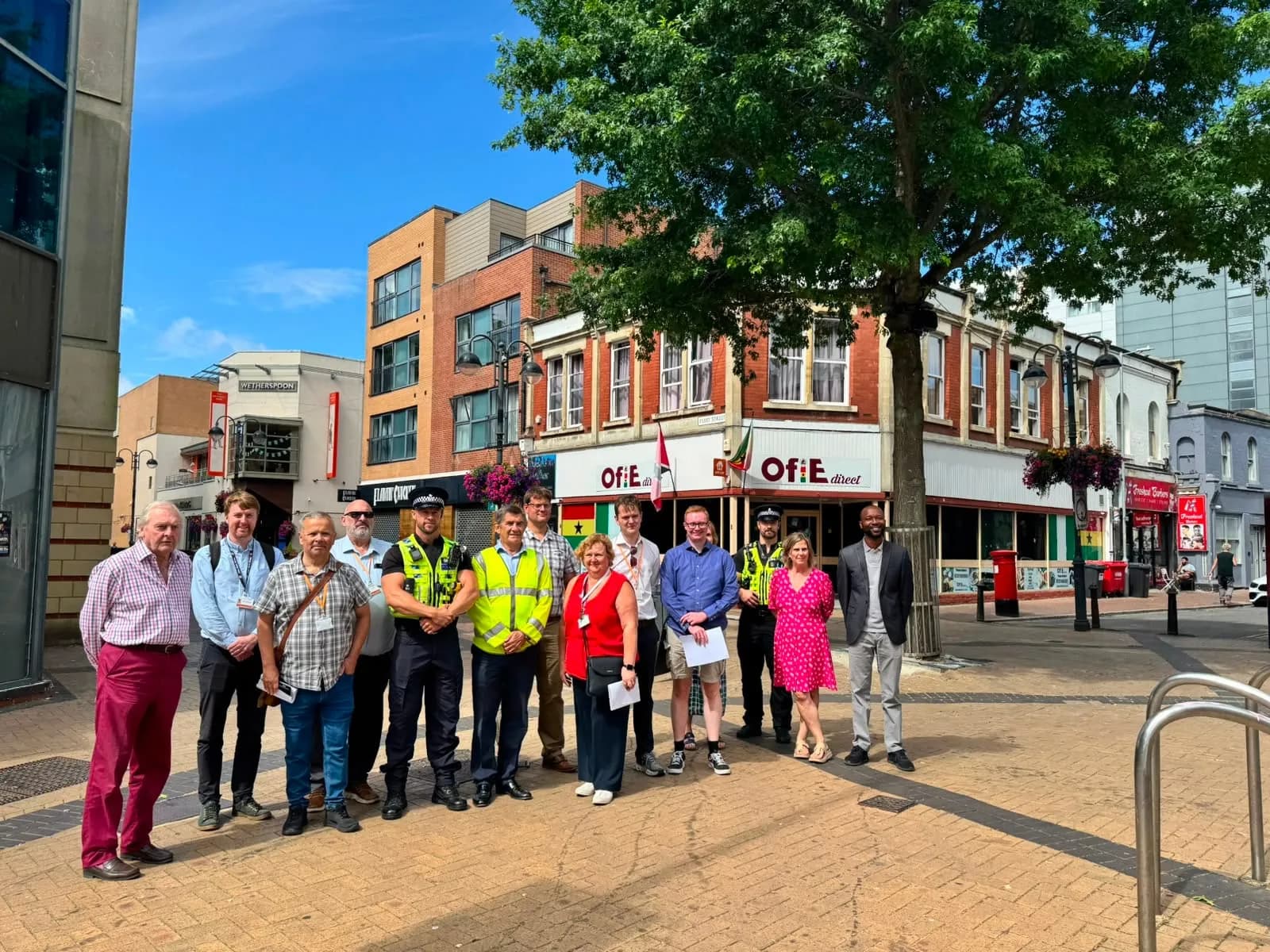 A group of diverse people, including police officers, stand on a paved street in front of shops and buildings under a bright sky with a large tree.