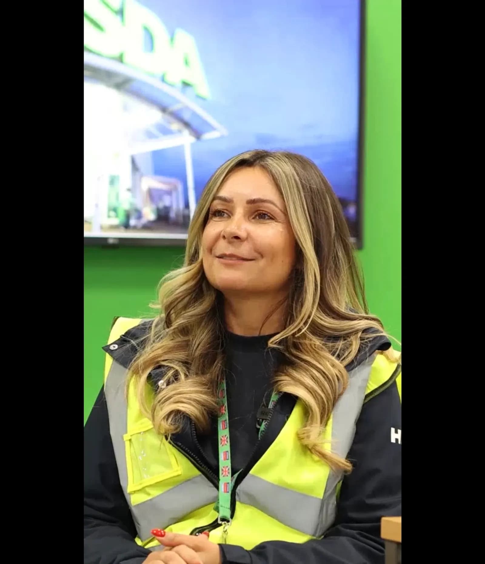 A woman with long wavy blonde hair and a dark shirt under a reflective safety vest smiles, with a large screen displaying a building and the word "ASDA" behind her.