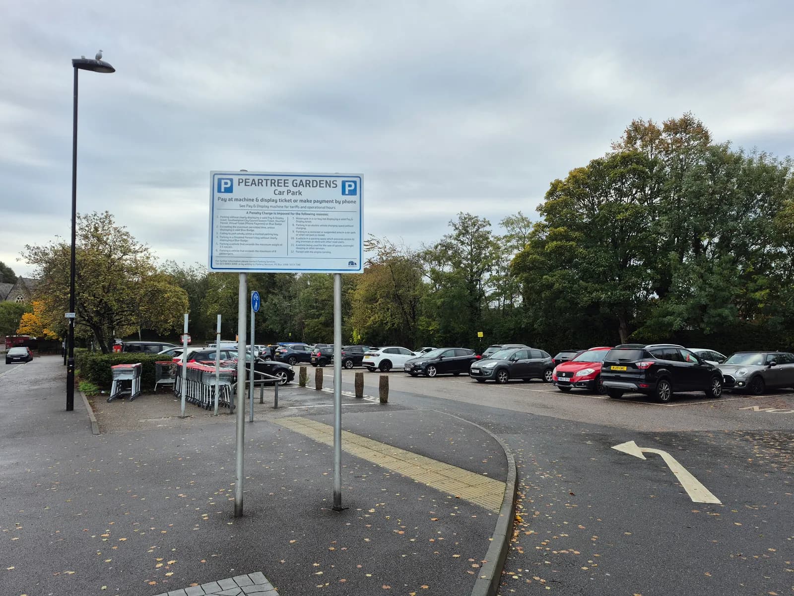 A car park with a large sign for "PEARTREE GARDENS Car Park" in the foreground, showing numerous cars parked under a cloudy sky.