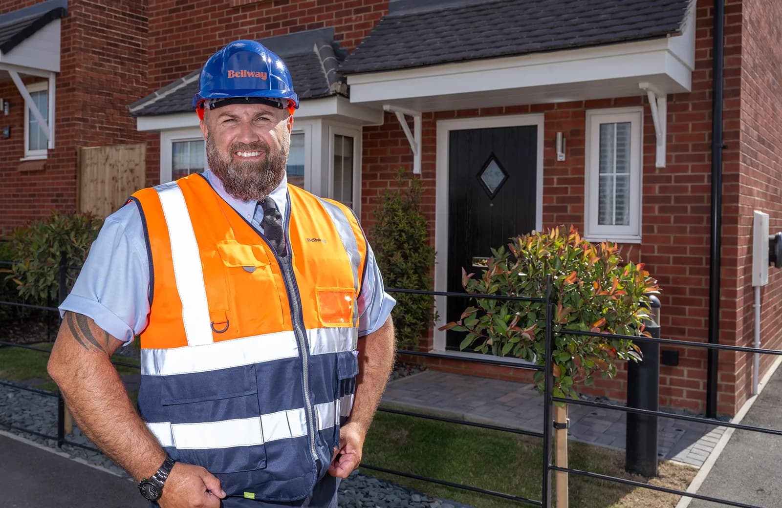 A smiling man wearing a blue hard hat, hi-vis vest, and shirt stands in front of a modern brick house with a black front door.