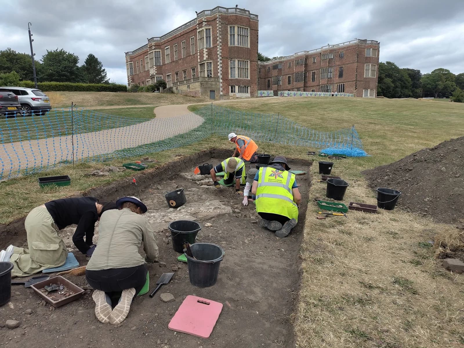 Archaeologists in an excavation trench carefully uncover artifacts with tools, with a large historic brick building and green lawn in the background under a cloudy sky.