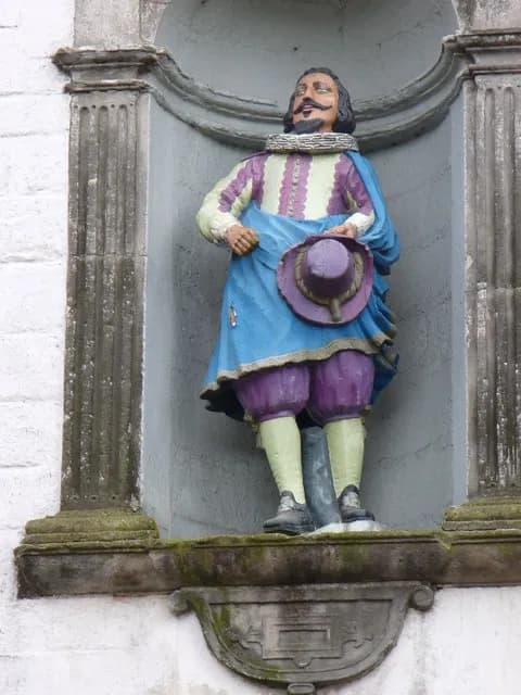 A colorful statue of a man with a mustache, wearing period clothing and holding a purple hat, stands in a stone niche.