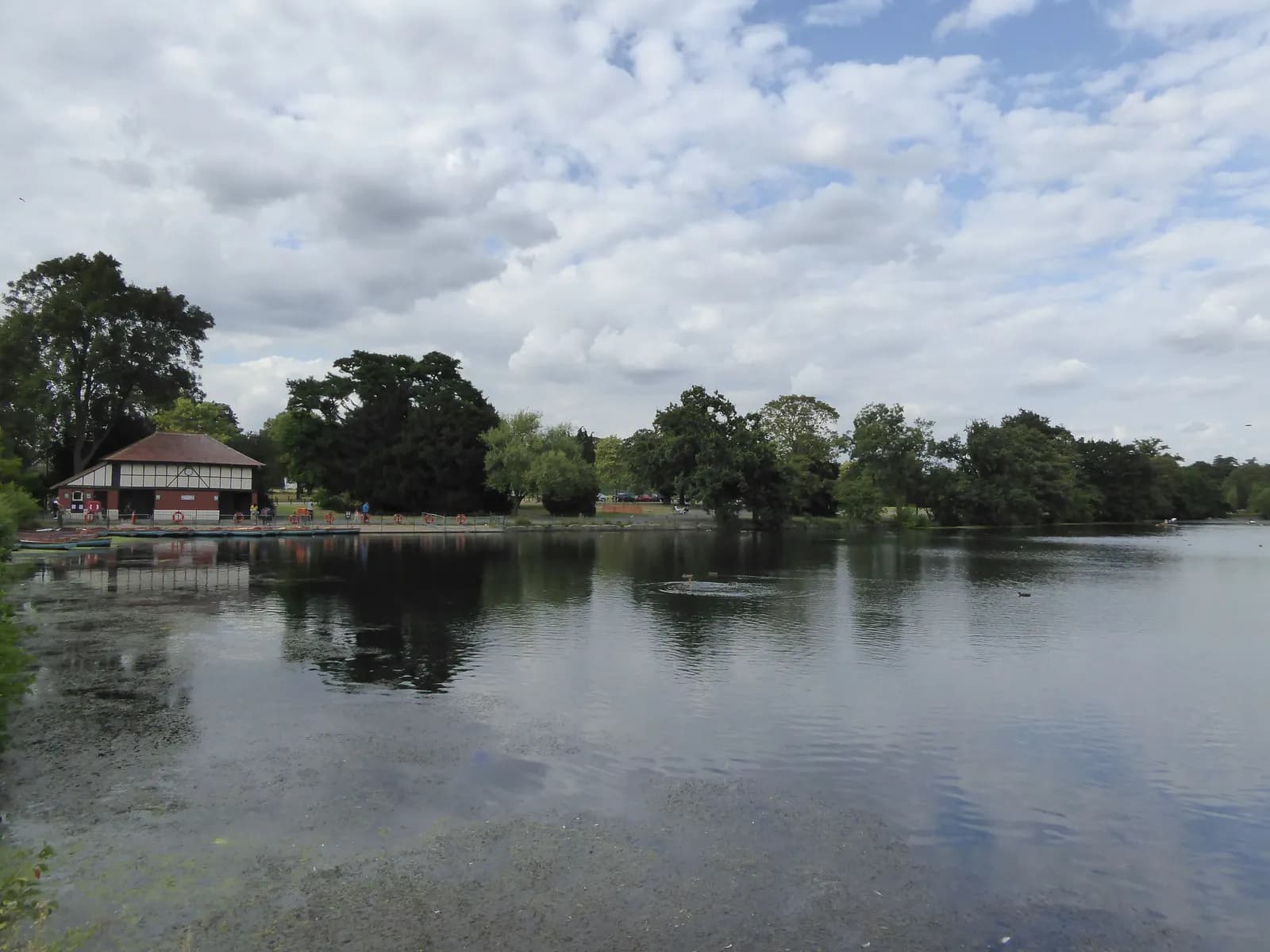 A red and white boathouse with a brown roof sits on the edge of a calm lake under a cloudy sky, surrounded by lush green trees.