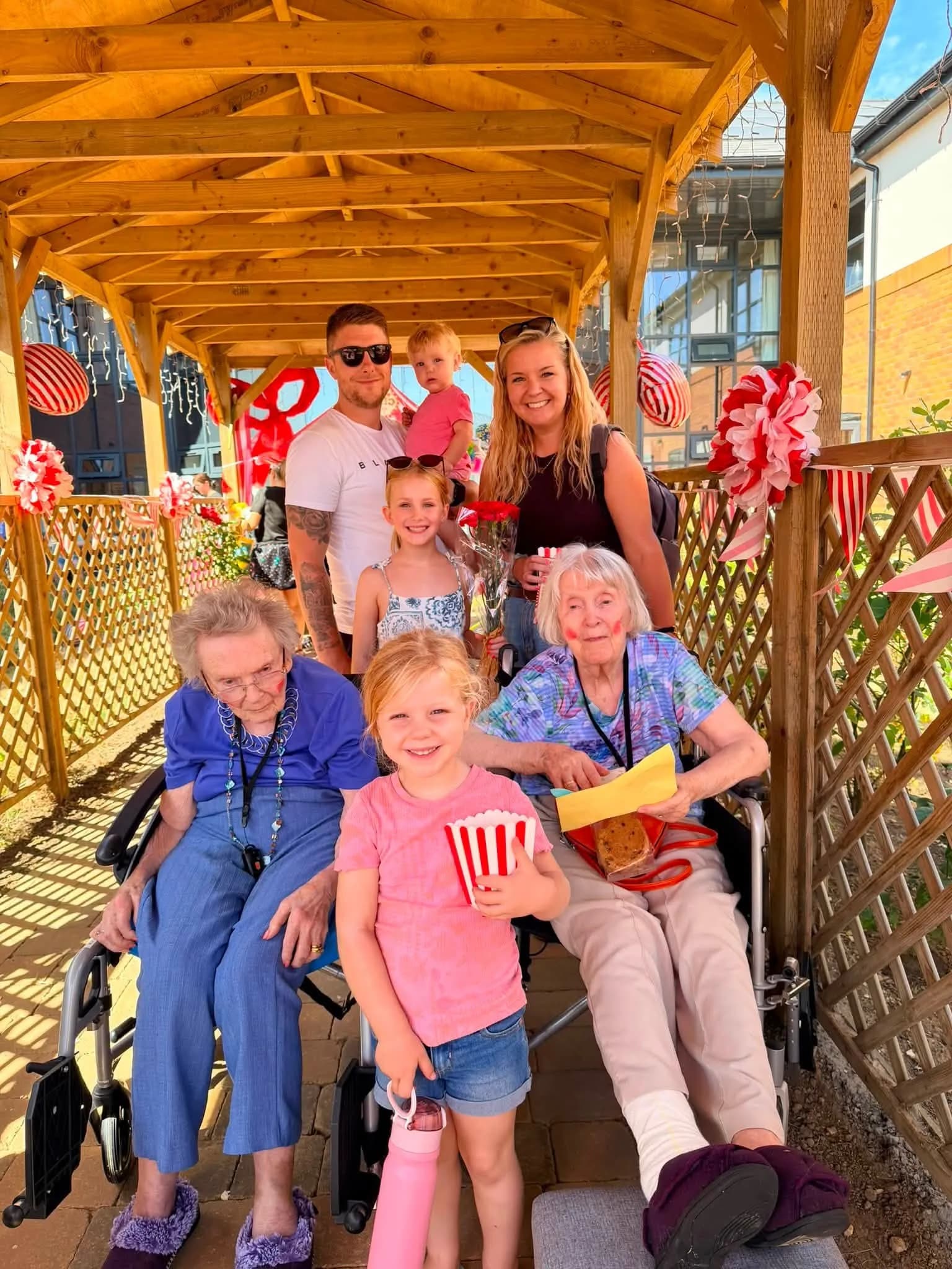A family portrait shows three generations gathered under a wooden pergola, with two elderly women in wheelchairs in the foreground, and a young couple with two children standing behind them, all smiling.