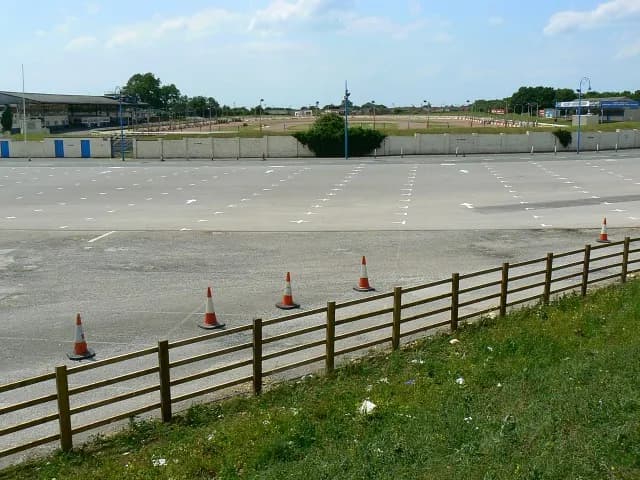 A wide shot of an empty concrete parking lot with painted white arrows and a wooden fence in the foreground against a backdrop of distant buildings and trees under a clear sky.