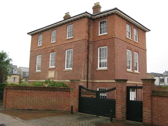 A large red brick house with white trim windows and dark green gates in the foreground, with other houses visible in the background.