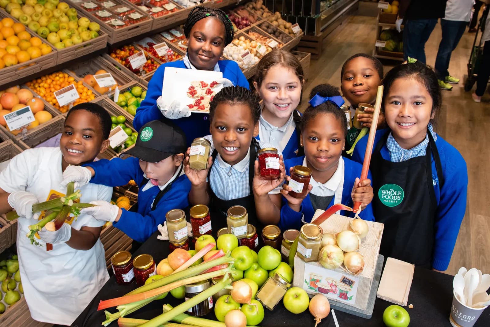 A group of smiling schoolchildren are proudly displaying jars of homemade preserves and fresh produce like apples, rhubarb, and onions at a market stall, with shelves of fruit and vegetables in the background.