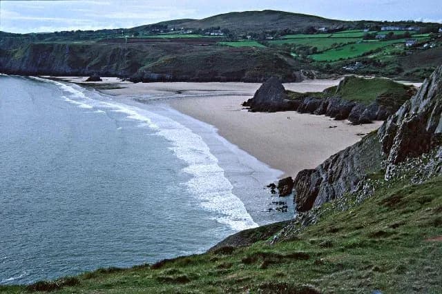 A scenic view of a sandy beach curving along the coast, with waves gently breaking on the shore and lush green hills rising in the background.