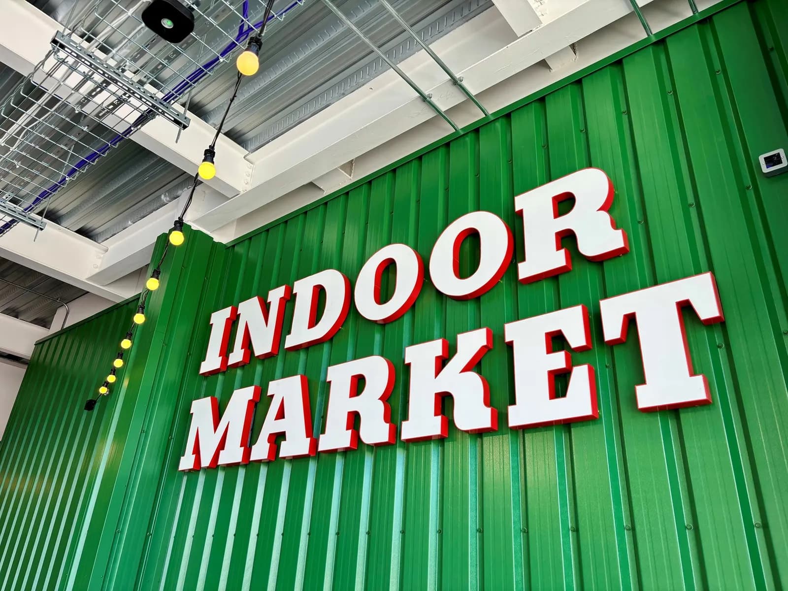 A large sign for an "INDOOR MARKET" in white letters with red borders on a corrugated green wall, with string lights hanging from the ceiling.