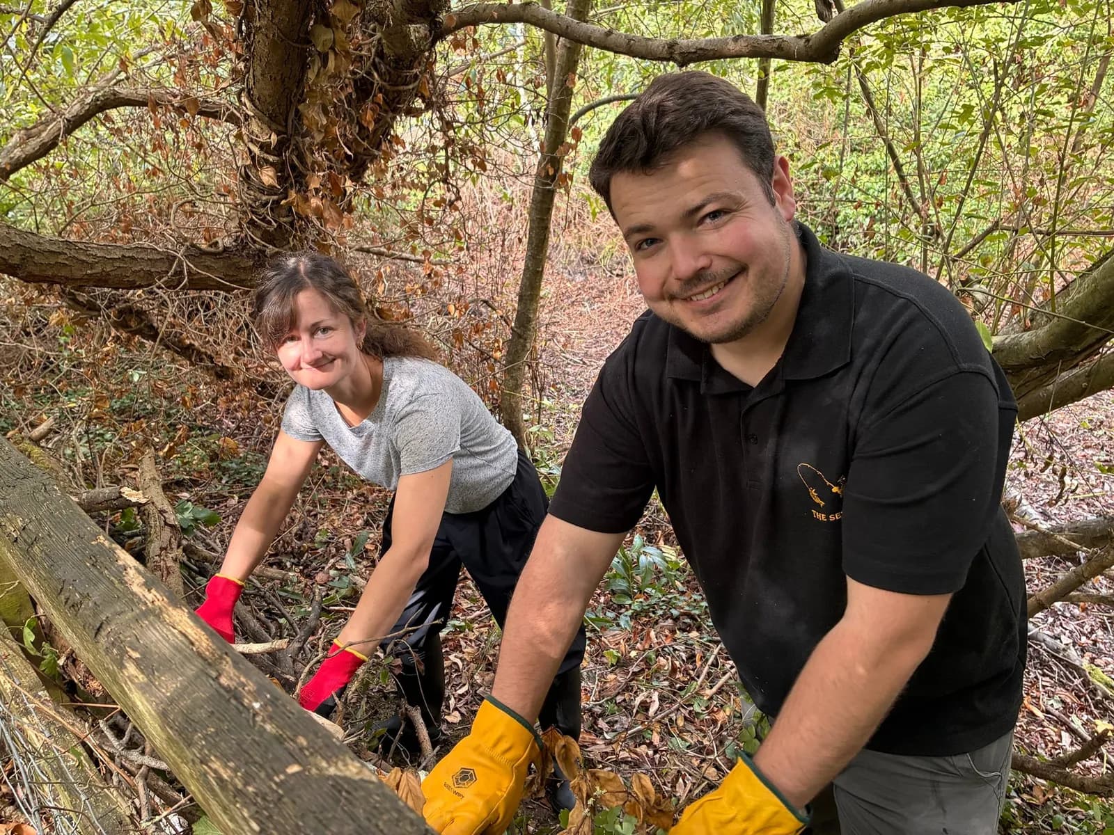 A man and a woman in work gloves smile as they clear brush and fallen branches in a wooded area.