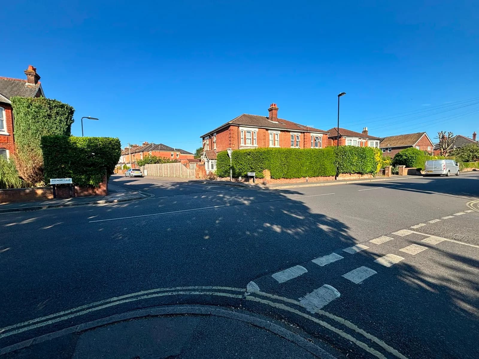 A quiet residential street intersection with traditional brick houses partially obscured by tall hedges, under a clear blue sky.