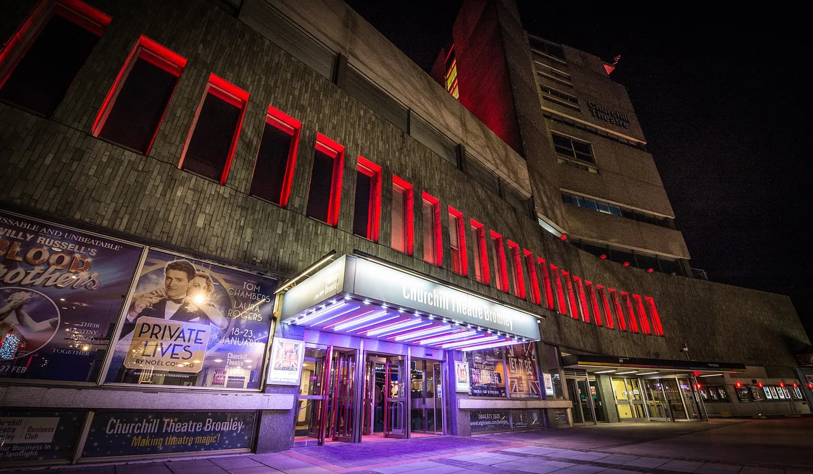 The Churchill Theatre Bromley at night, with its entrance canopy brilliantly lit in purple and red windows glowing on the concrete facade.