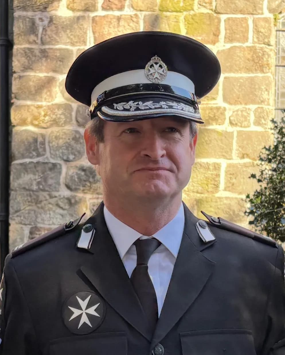 A man in a black uniform with a decorative cap and a white cross emblem on his chest, standing against a stone wall.