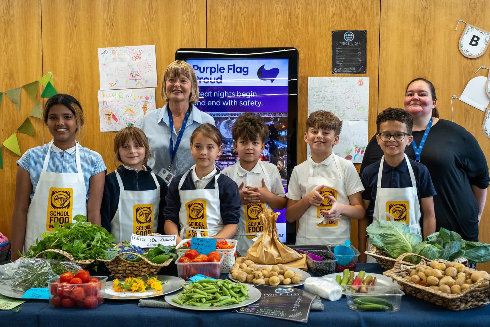 A group of cheerful children and two adults standing behind a blue table laden with fresh vegetables, fruit, and baked goods, with a digital display in the background.