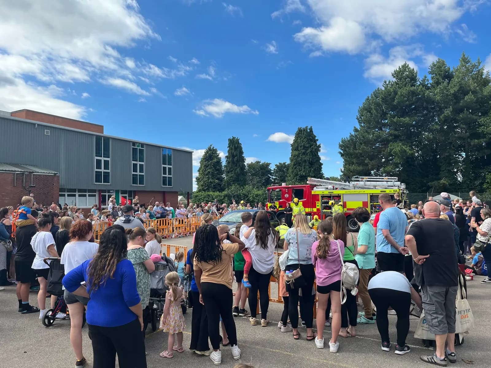 A crowd of people, including many children and adults, stand outdoors watching a red fire truck with several firefighters in yellow high-visibility jackets.
