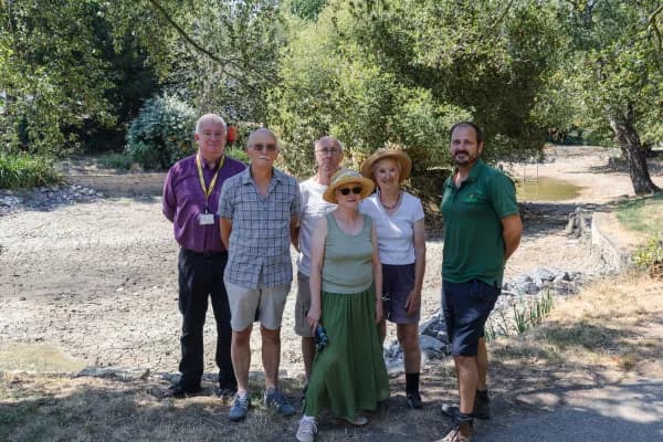 Six adults stand together, two men on the left, a man and two women in the middle, and one man on the right, in front of a dried-up riverbed under a sunny, treed sky.