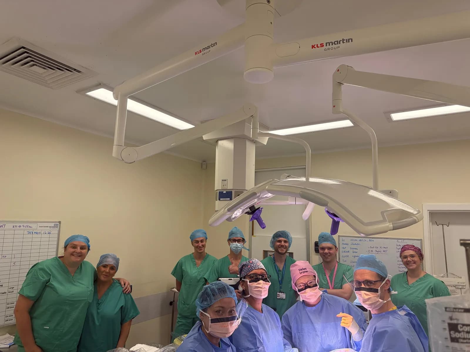 A team of medical professionals in scrubs and masks stand together in an operating room with surgical lights overhead.