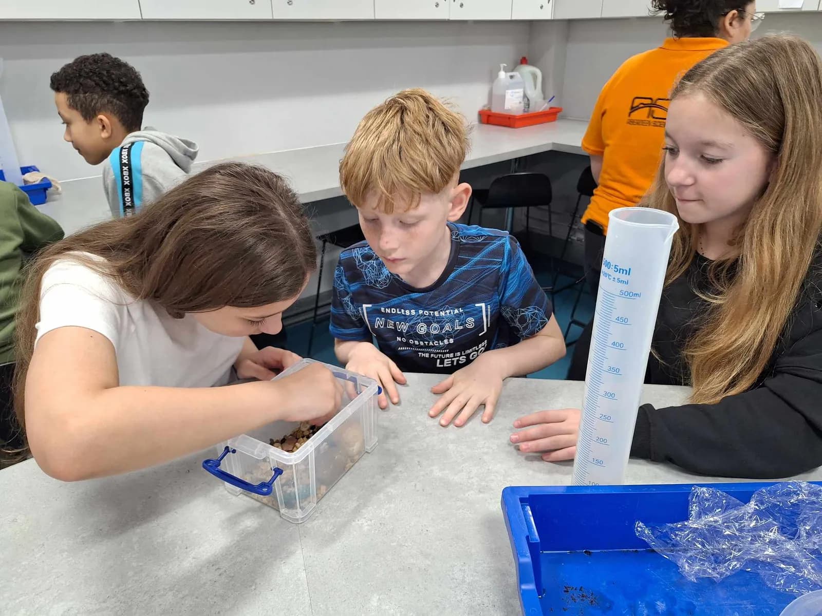Three children are gathered around a table with various science equipment, one girl is looking into a container while another girl holds a measuring cylinder.