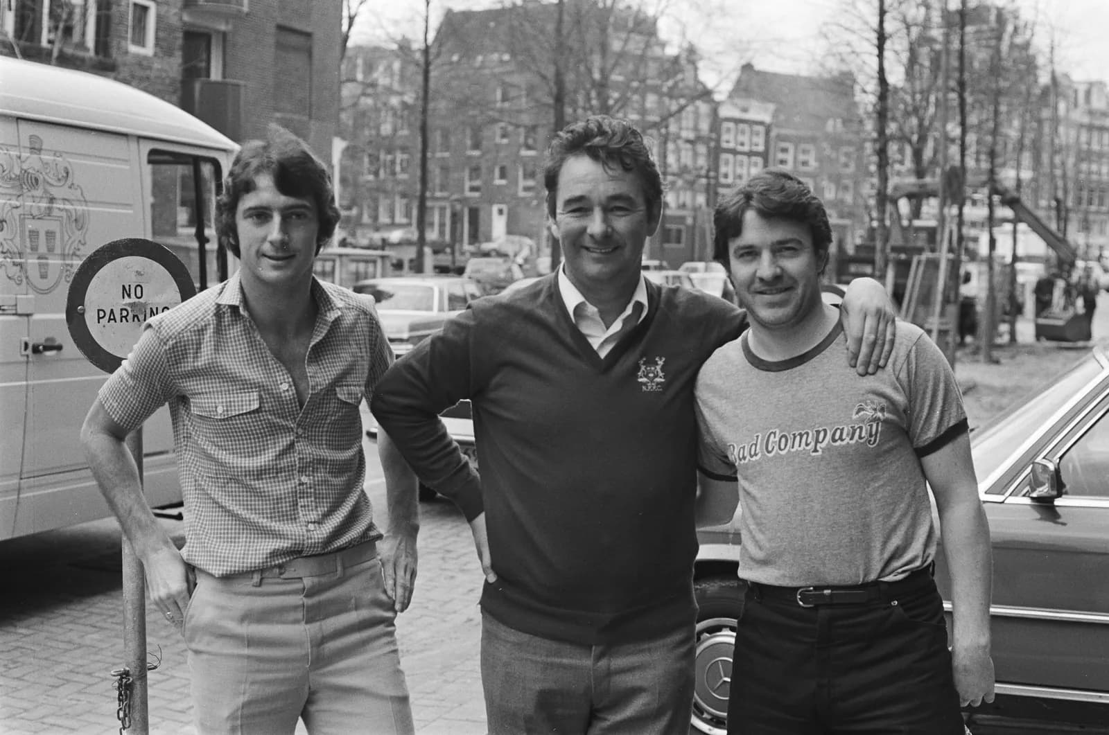 Three men stand together smiling at the camera outdoors, with buildings and a car in the background.