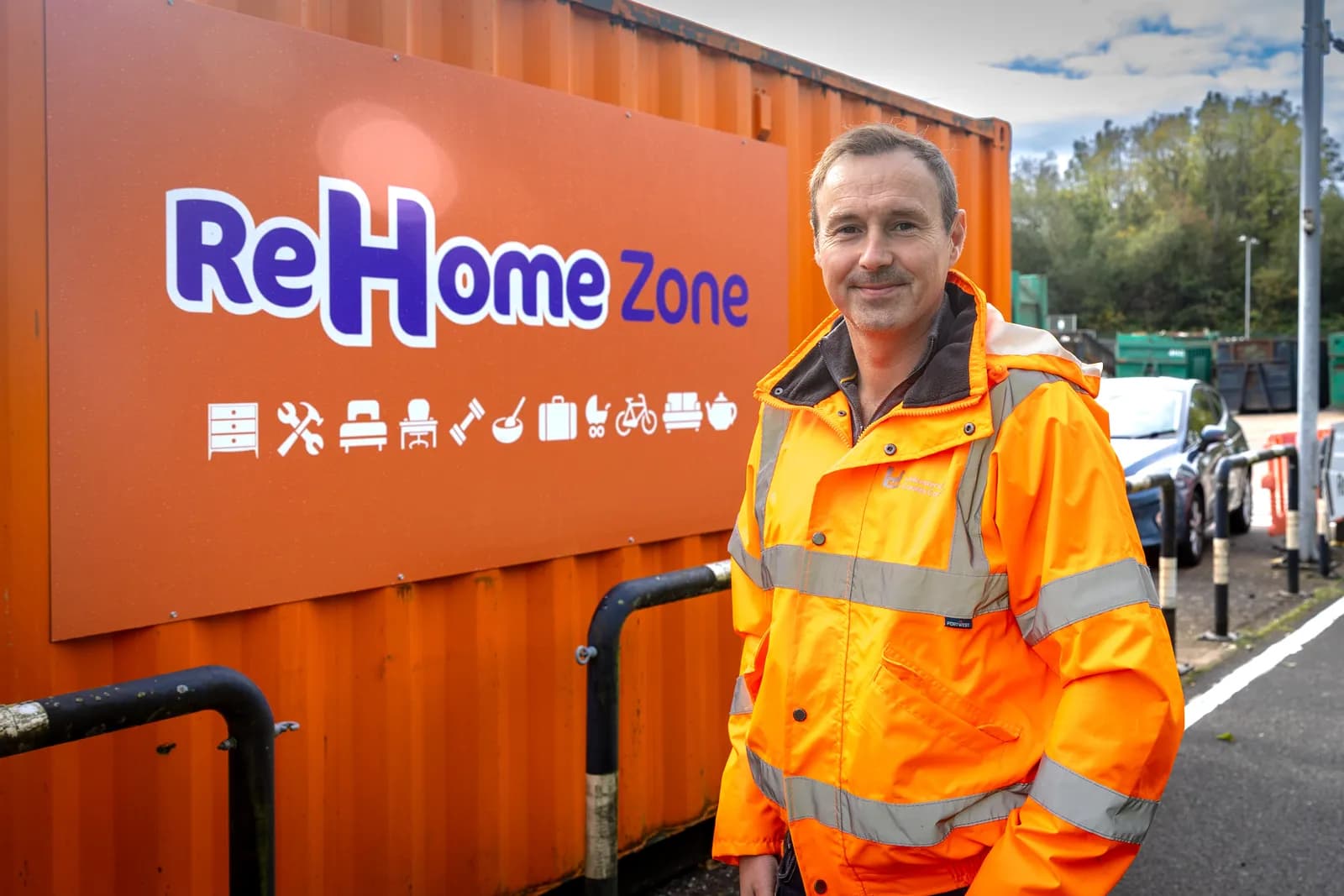 A man in an orange high-visibility jacket stands smiling in front of a rust-colored container with a white and purple ReHome Zone logo.