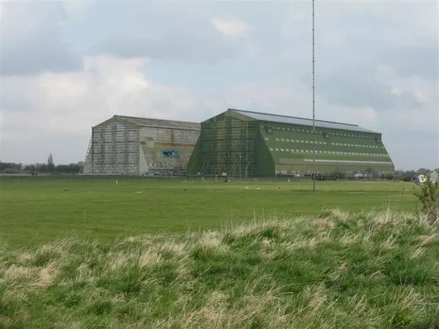 Two large, derelict aircraft hangars stand in a vast green field under a cloudy sky.