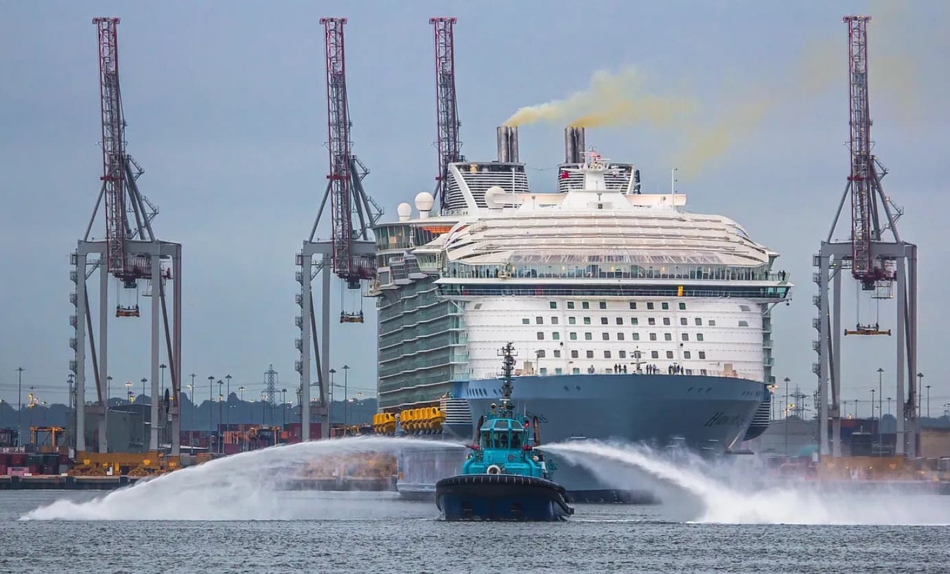 A tugboat sprays water in front of a large cruise ship in a harbor with port cranes in the background.