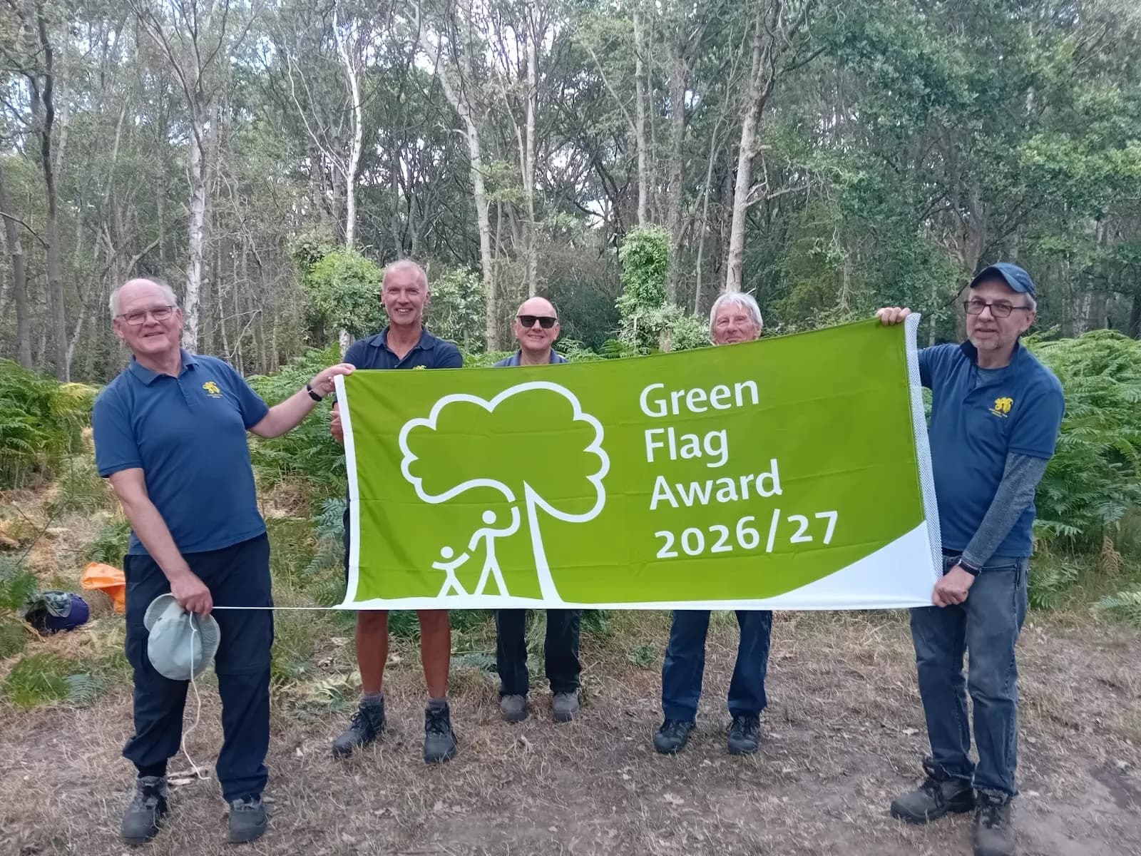 A group of five men stand in a forest clearing, smiling and holding a green banner that reads "Green Flag Award 2026/27" with a white tree and people logo.