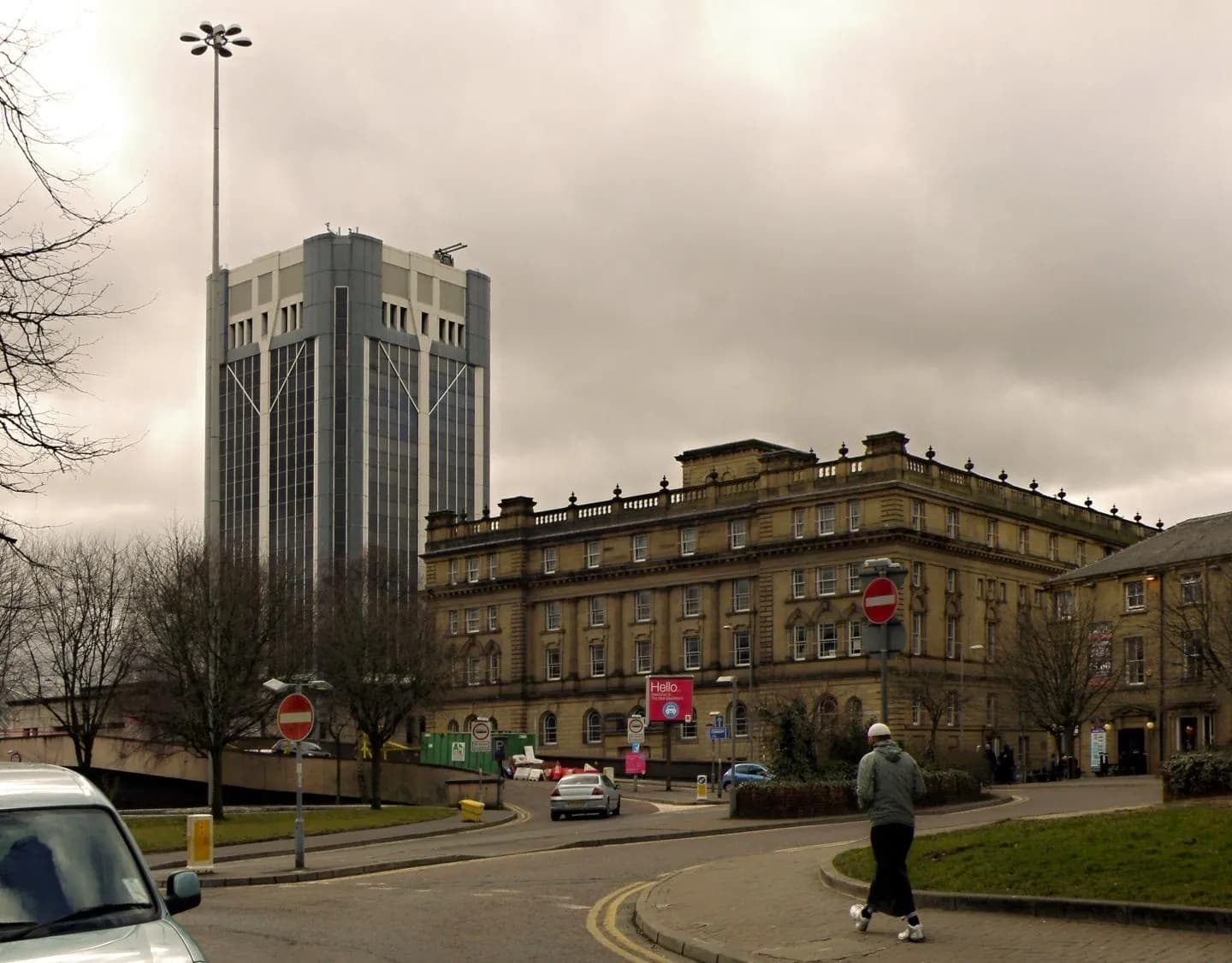 A tall, modern building with a large street light at its top, stands behind a classic stone building next to a curved road where a person walks.