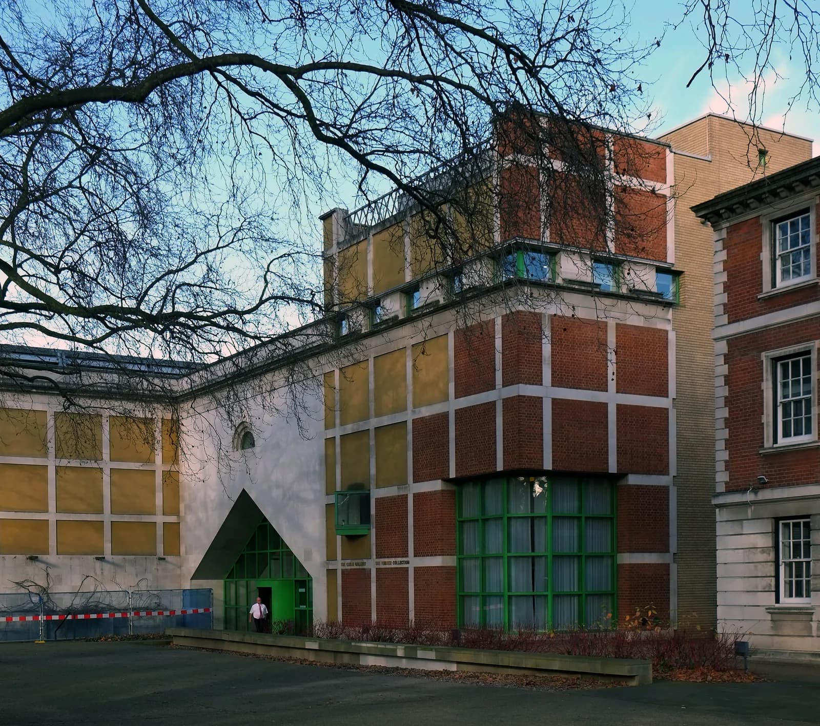 A modern building with a stark white, triangular entrance and grid-patterned brown brickwork, sits under the sparse branches of a winter tree.