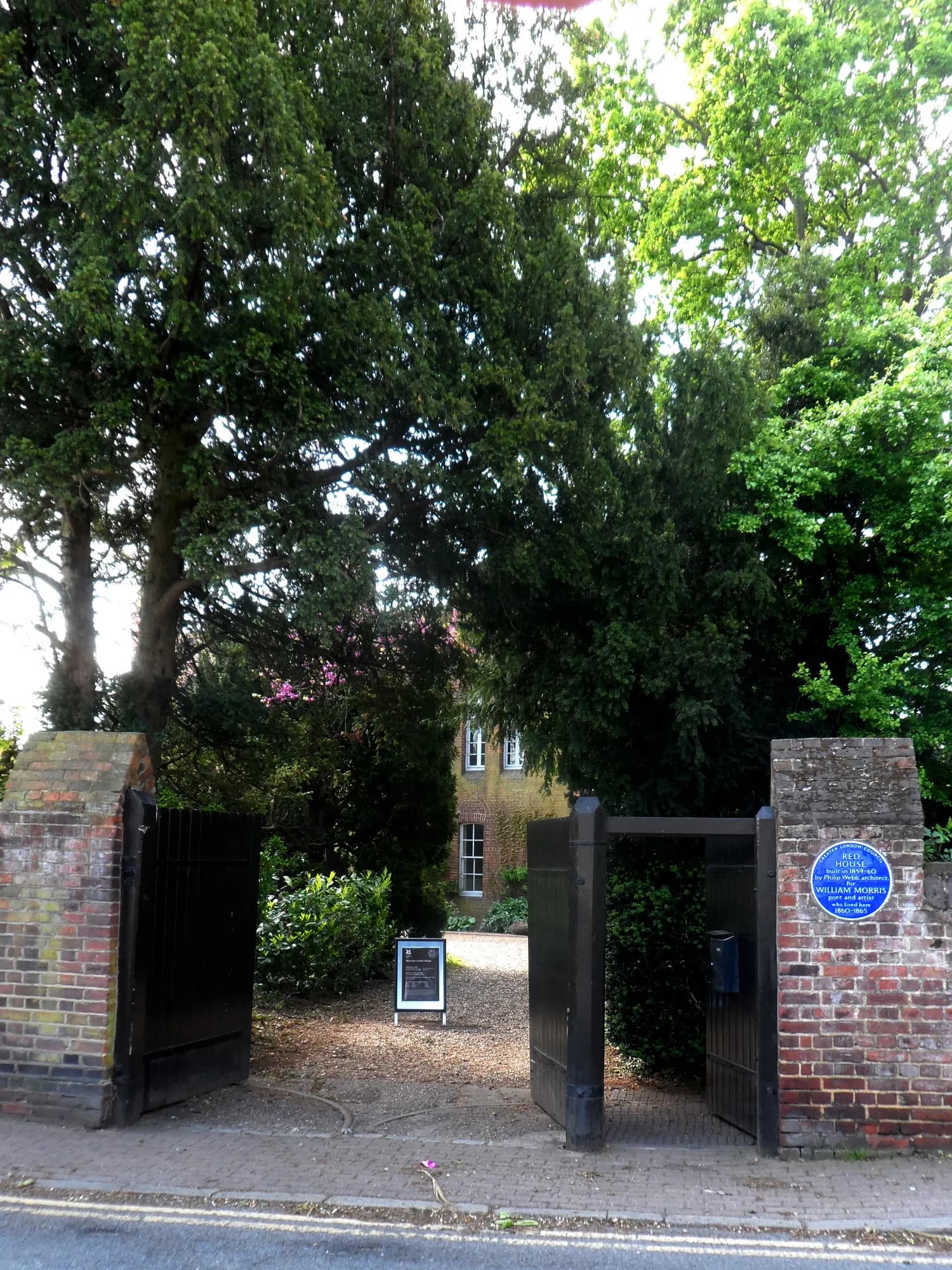 A driveway entrance with open black gates leads to a historic building partially visible through lush green trees, with a blue plaque on the right brick pillar.