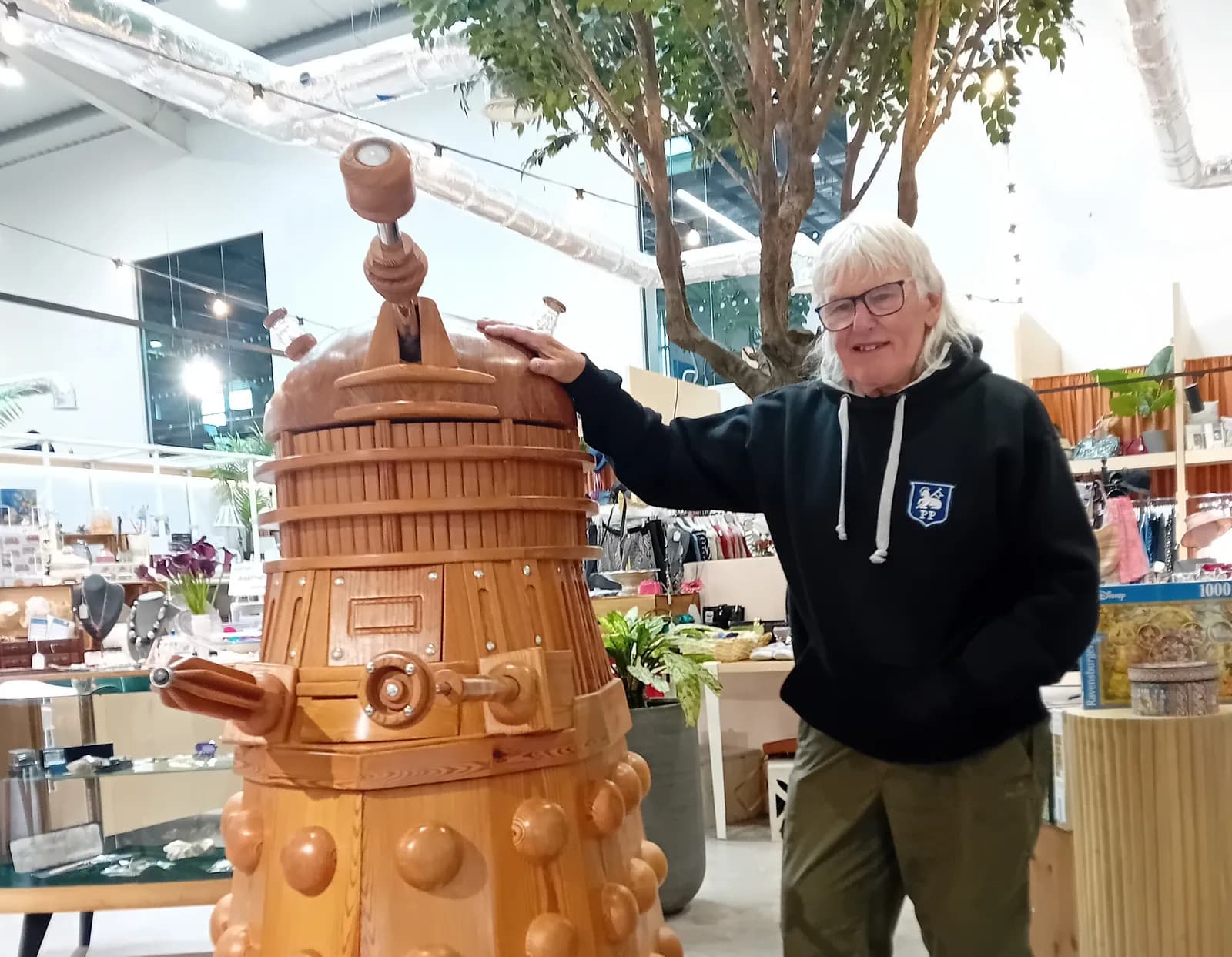 A person with white hair and glasses, wearing a black hoodie and olive green pants, stands next to a large wooden Dalek.