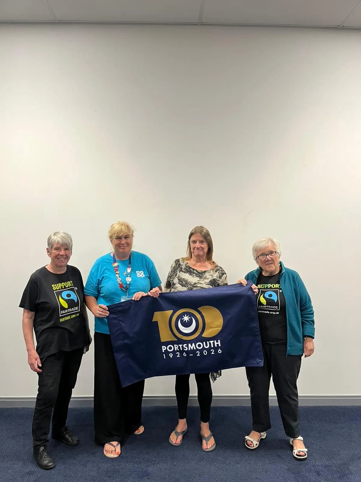 Four women holding a blue banner that reads "100 Portsmouth 1926-2026" in front of a white wall.