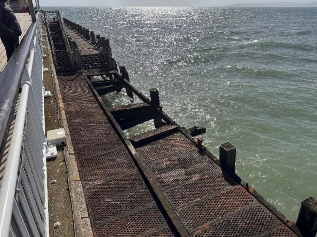 A long, rusted metal walkway extends into shimmering water, supported by old wooden structures, with a railing visible on the left.