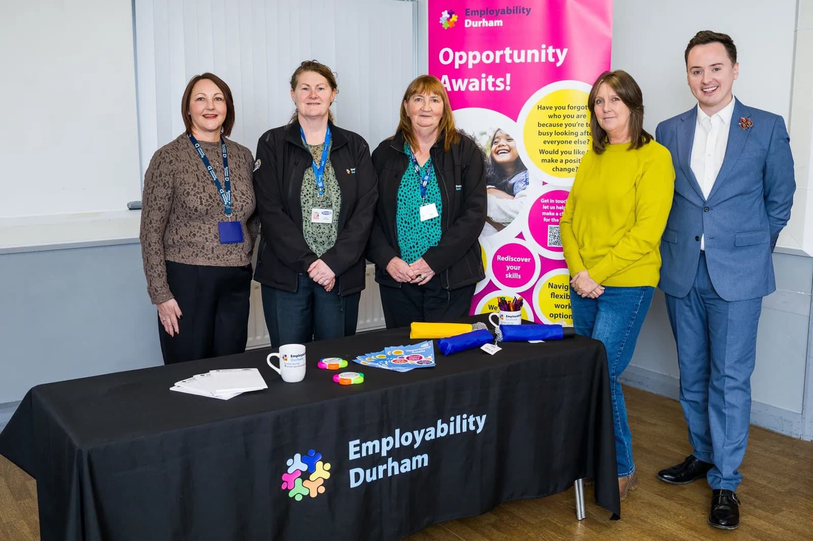 Five people, four women and one man, stand behind a black table with a sign for Employability Durham, and a pink banner is visible behind them.