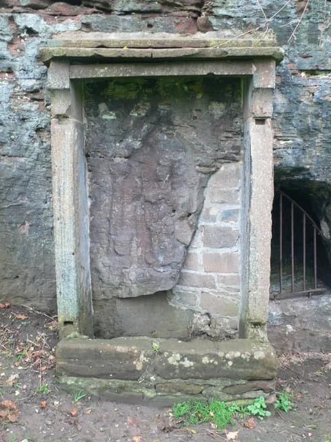 A weather-beaten stone structure with an open archway containing a rough stone and brick wall, set against a background of natural rock and a dark opening with iron bars.