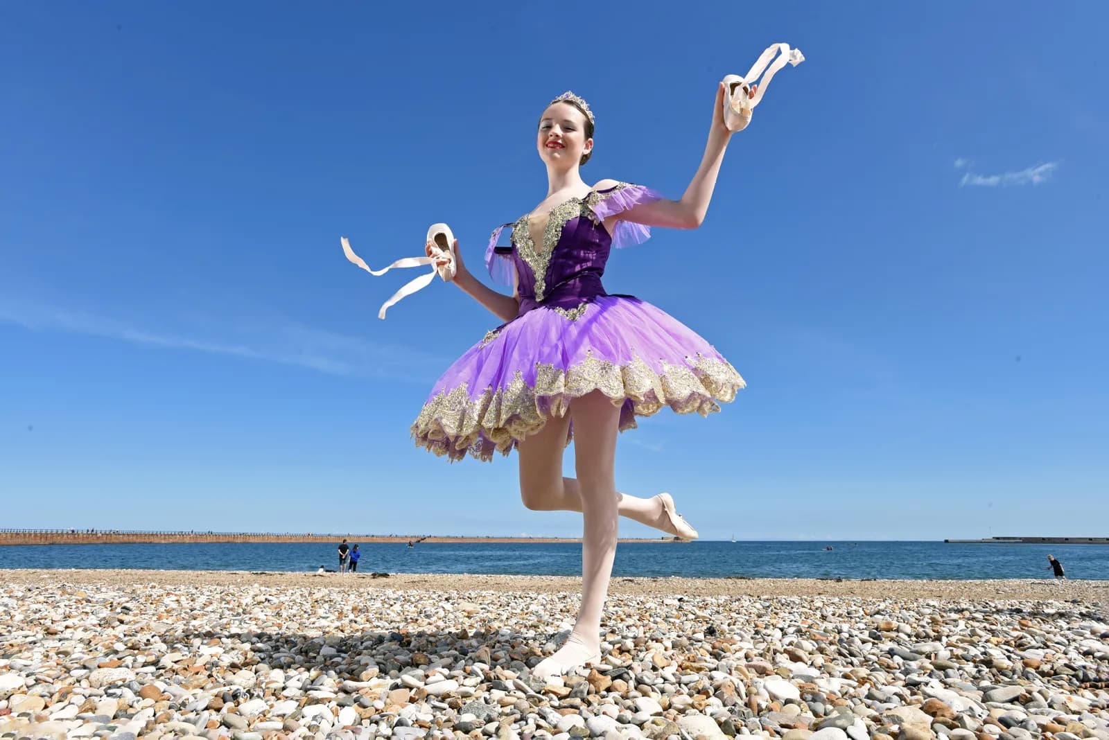 A young ballet dancer in a purple tutu smiles while holding ballet shoes and posing on a pebble beach with the sea and a clear blue sky in the background.