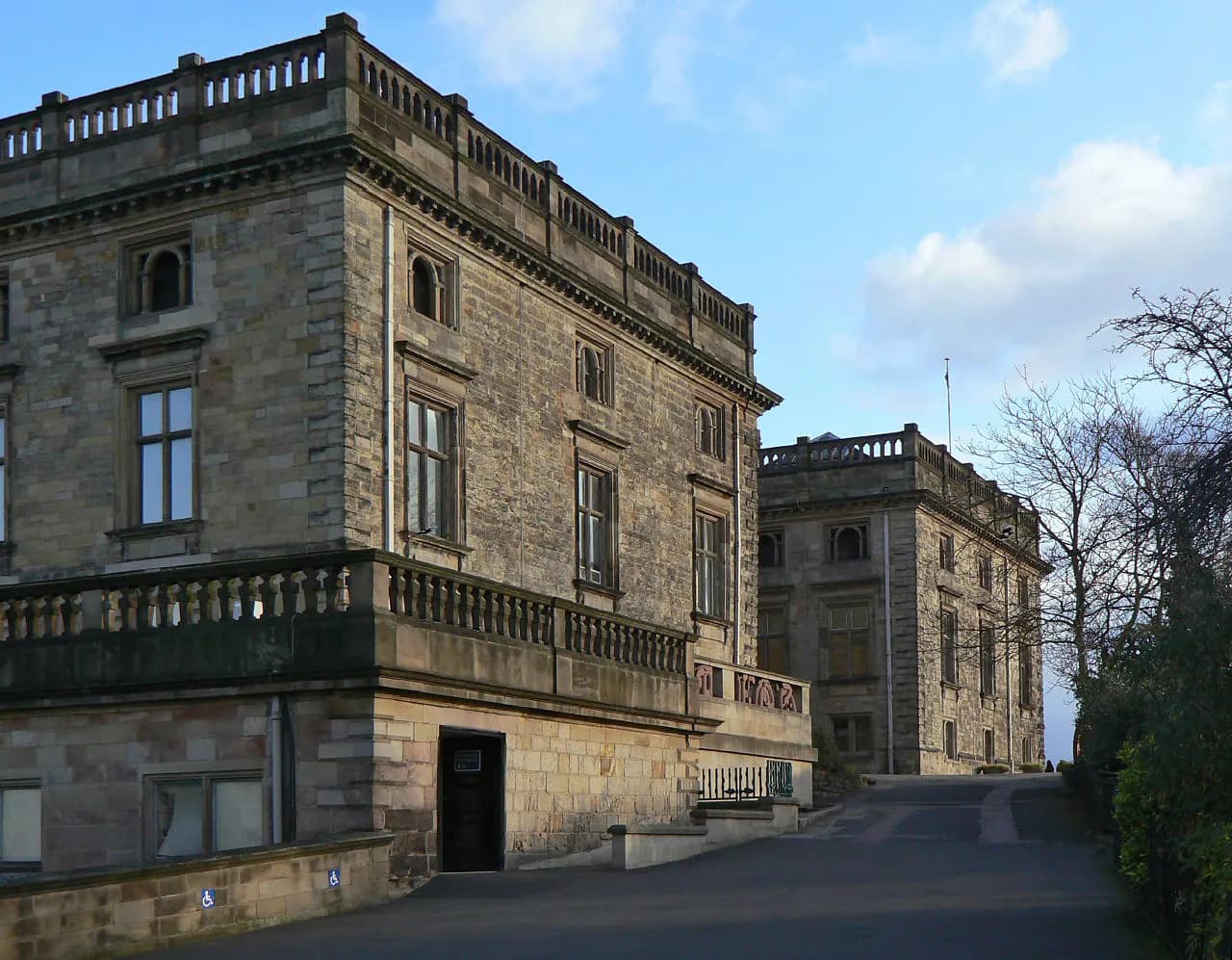 A large stone building with multiple windows and a railing-lined balcony, with a road leading up to a second, similar building in the background.
