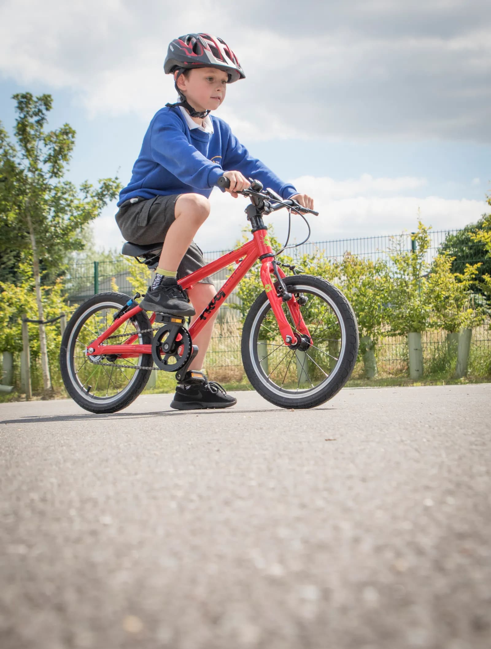 A young boy in a blue long-sleeved shirt, shorts, and a helmet rides a red bicycle on an asphalt path with green trees and a fence in the background under a cloudy sky.