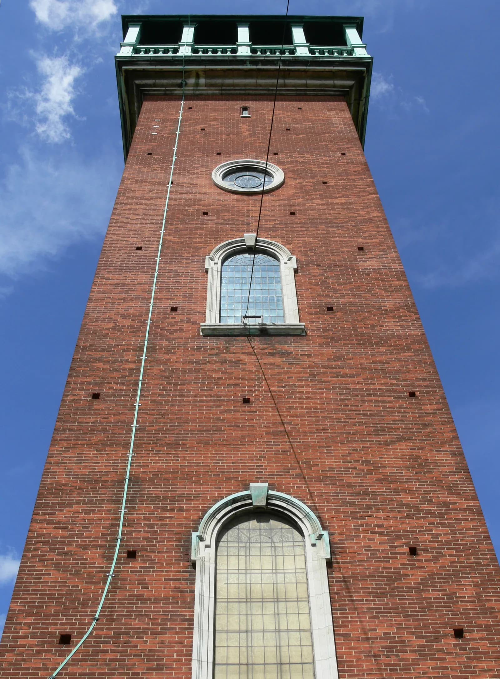 A low-angle view of a tall rectangular brick tower with a green balcony and arched windows against a blue sky with some clouds.