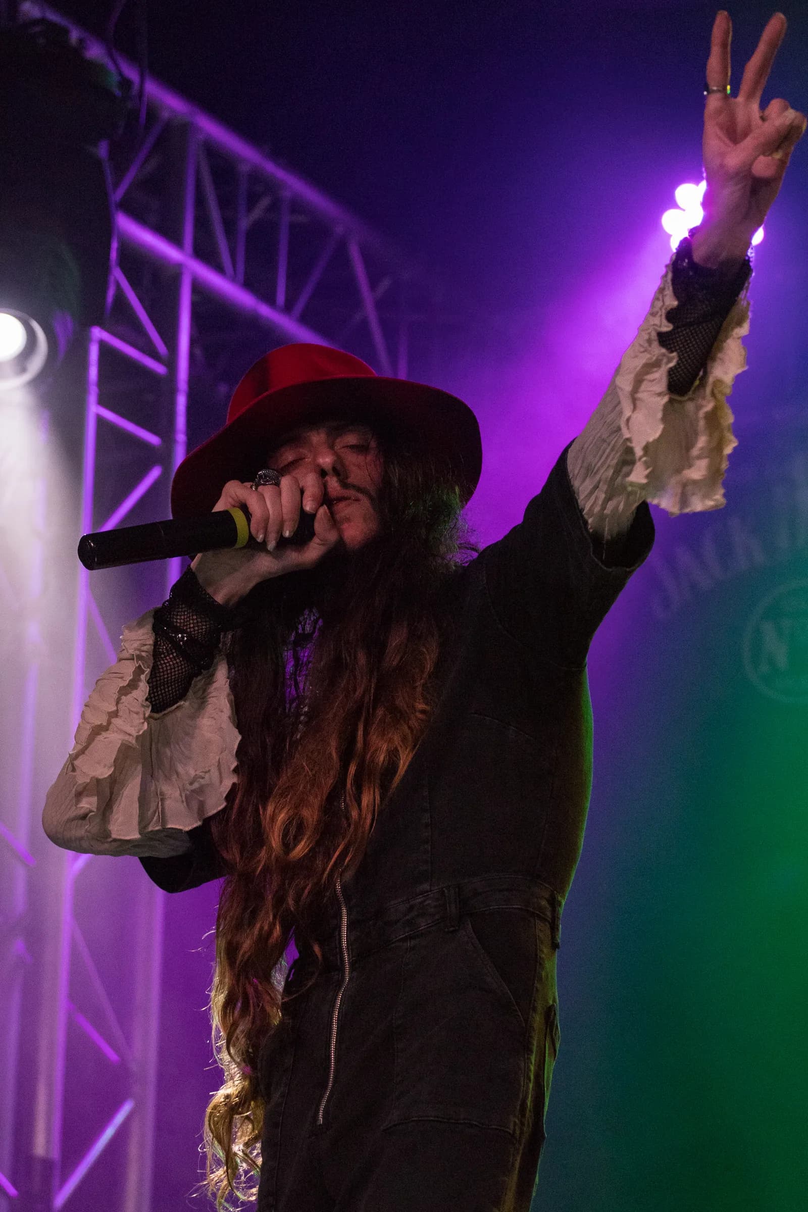 A singer with long hair and a red hat, holding a microphone while performing on stage with purple and green lighting, raises his left hand making the peace sign.