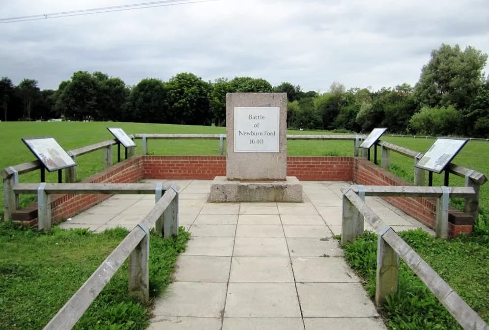 A historical marker for the Battle of Newburn Ford 1640 stands on a paved area surrounded by short brick walls and wooden fences, with green grass and trees in the background under a cloudy sky.